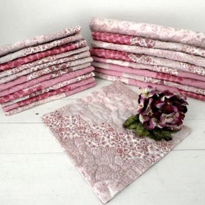 May include: Several stacks of folded pink and white patterned quilts. The quilts feature floral and geometric designs. An unfolded quilt is displayed with a bouquet of dark red flowers. The quilts are on a white surface.