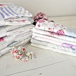 May include: Two stacks of folded quilted fabric pieces featuring various floral and striped patterns in shades of pink, blue, and white. Each stack is tied with a white ribbon and decorated with a small cluster of artificial pink flowers.