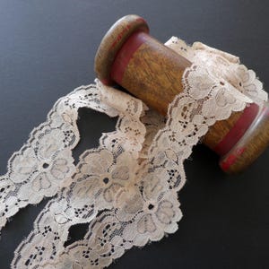 May include: A wooden spool with pale peach lace. The lace has a floral design and is wrapped around a wooden spool with red bands. The background is a dark colour, which makes the lace stand out. The lace is a delicate, vintage style.