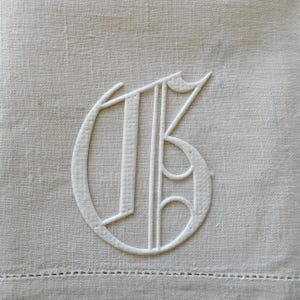 May include: A light beige linen napkin featuring a white embroidered monogram of the letter G in a gothic script style. The fabric has a visible woven texture and a decorative hemstitch detail along the bottom edge of the square textile piece.