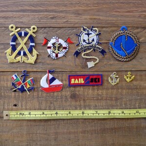 May include: A collection of nine nautical themed iron-on patches. The patches feature anchors, life preservers, sailboats, and the text "Sail & Ski".