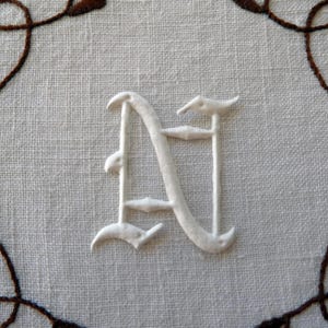 May include: A white linen fabric square featuring a raised, white embroidered letter N in a gothic-style font. The letter is centred on the fabric, which is framed by a dark brown embroidered border with curved, decorative line patterns.