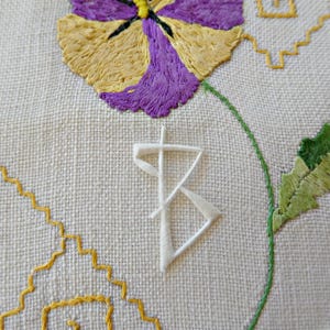 May include: A piece of off-white fabric featuring hand embroidery of a purple and yellow pansy flower, a green stem, and a yellow geometric border. A white, stylised letter B is embroidered in the centre of the fabric, layered over the design.