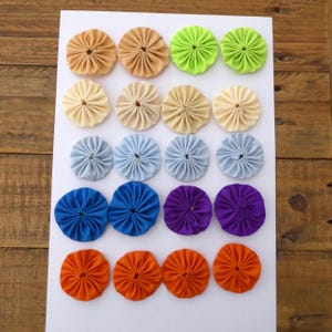 May include: Sixteen fabric rosettes in a variety of colours, arranged in four rows. Colours include tan, light green, cream, light blue, royal blue, purple, and orange. The rosettes are made of pleated fabric and are circular in shape.
