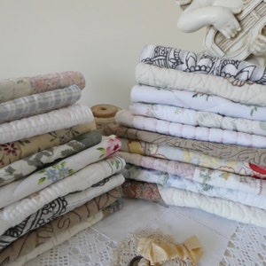 May include: A stack of vintage fabric scraps in various patterns and colors, including floral, paisley, and plaid. The fabrics are mostly white, cream, and beige, with some pink and blue accents. The fabrics are arranged in a pile on a white lace tablecloth.