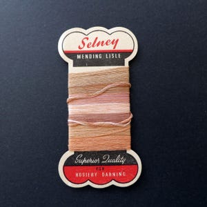Peut inclure: Une bobine en carton vintage enroulée de fil à repriser en fil d'Écosse dans des tons beige, sable et brun clair. La bobine porte le nom de marque Selney, ainsi que les inscriptions Mending Lisle, Superior Quality et For Hosiery Darning.