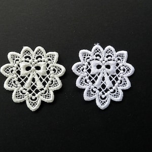 May include: Two circular lace appliqués featuring a central bow motif surrounded by a scalloped, openwork floral pattern. One appliqué is off-white, and the other is bright white, both showcasing intricate needlework and delicate thread detailing.