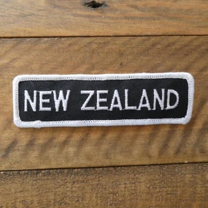 May include: A rectangular embroidered patch with a black background and white border. The text NEW ZEALAND is stitched in white capital letters across the centre of the patch. The design features a clean, bold font with a finished stitched edge.