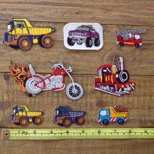 May include: A selection of embroidered vehicle patches, including a yellow and blue dumper truck, a purple off-road vehicle, a red fire engine, a red motorcycle with flames, and a red train. Patches are displayed on a wooden surface.