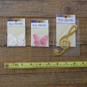 May include: Three iron-on patches on white card stock packaging. The patches are a white butterfly, a pink butterfly, and a gold treble clef. The packaging has the text "Kay Motifs" printed on it.
