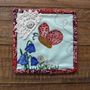May include: A handmade square quilt block featuring a blue and red butterfly, bluebell flowers, and a white crocheted doily. The block is edged with a red, white and blue patterned border.