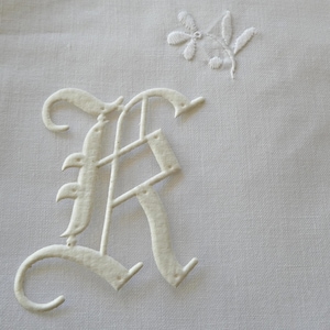 Peut inclure: Une surface en tissu blanc présentant une grande lettre K brodée en relief dans un style gothique. Au-dessus de la lettre, se trouve un petit brin floral délicatement brodé avec des feuilles et des bourgeons, le tout en fil blanc.