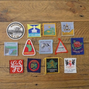 May include: A selection of vintage patches, featuring diverse designs and text, such as city and district names. The patches, in various shapes and colours, are arranged on a wooden surface.