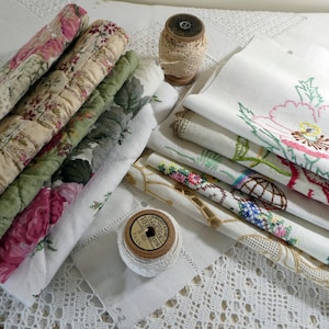 May include: A stack of vintage fabric scraps with floral embroidery and lace trim. The fabric scraps are arranged in a pile with the top layer folded over. There is a spool of white thread and a spool of white lace on the table.