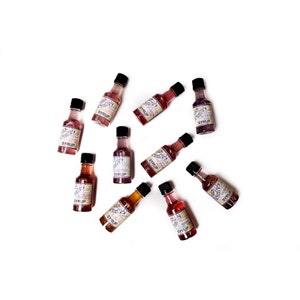 May include: Ten small glass bottles with labels that say "SYRUP" in a circle on a white background. The bottles are filled with a pink liquid.
