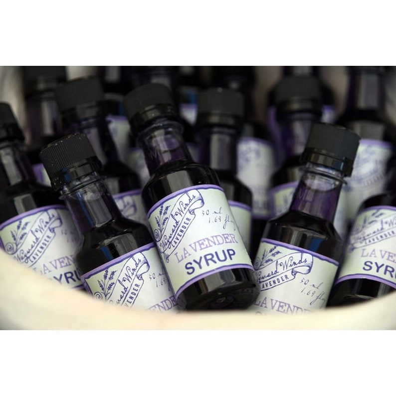 May include: A collection of small glass bottles filled with a dark purple liquid. Each bottle has a label that reads "Myward Winds Lavender Syrup" with a purple and white floral design. The bottles are arranged in a white ceramic bowl.