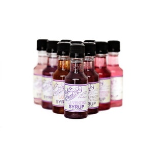 May include: A collection of eight small glass bottles with colorful labels. The bottles contain flavored syrups, including lavender, vanilla, and other flavors. The labels feature a purple and white design with the text "Whispered Winds" and "All Natural".