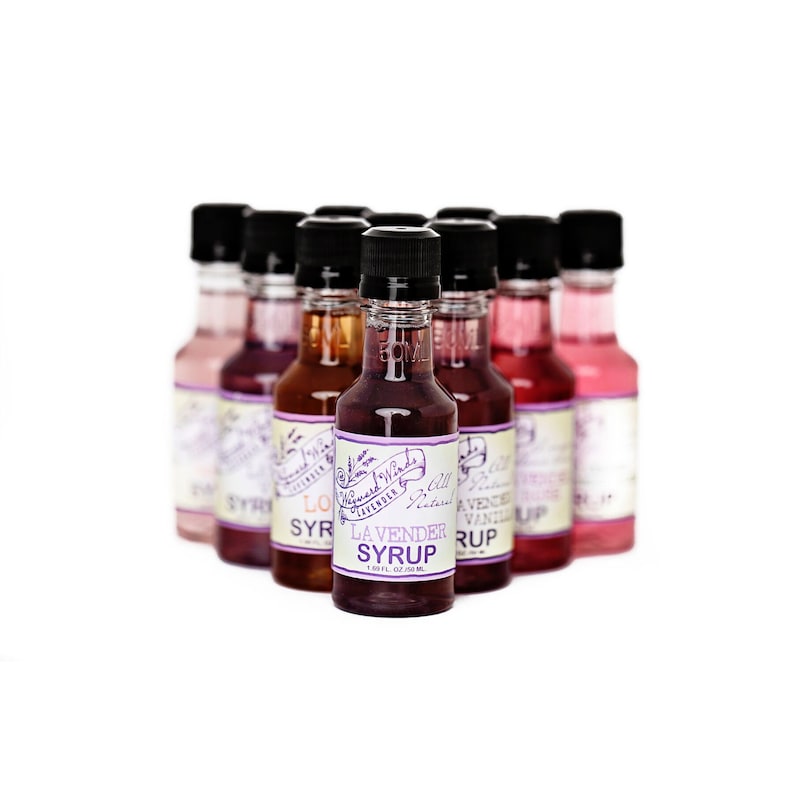 May include: A collection of eight small glass bottles with colorful labels. The bottles contain flavored syrups, including lavender, vanilla, and other flavors. The labels feature a purple and white design with the text "Whispered Winds" and "All Natural".