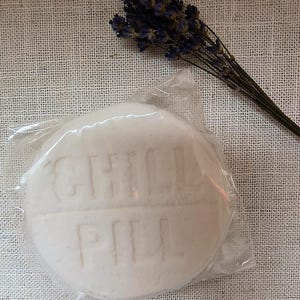 May include: A white round soap bar with the words "GINGER PEAR" embossed on the surface. The soap is wrapped in clear plastic and is sitting on a white fabric background.
