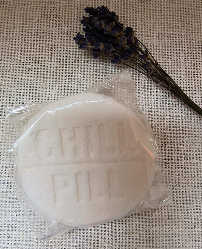 May include: A white round soap bar with the words "GINGER PEAR" embossed on the surface. The soap is wrapped in clear plastic and is sitting on a white fabric background.