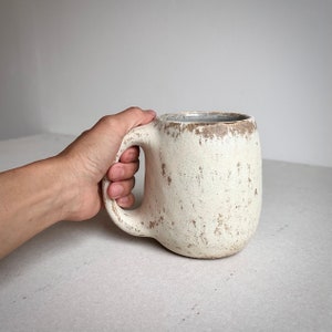 May include: A large, white ceramic mug with a speckled, rustic finish. The mug has a wide, rounded body and a large handle.