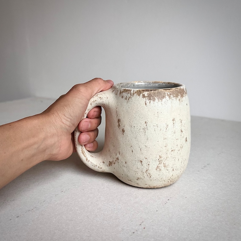 May include: A large, white ceramic mug with a speckled, rustic finish. The mug has a wide, rounded body and a large handle.