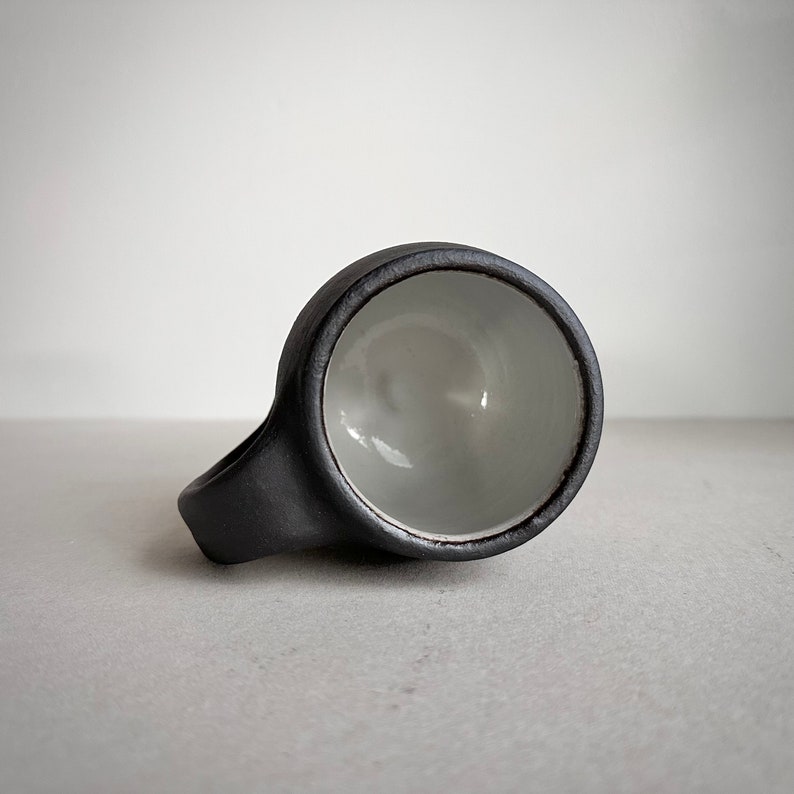 May include: A black ceramic mug with a white interior. The mug has a rounded handle and a wide, shallow bowl.