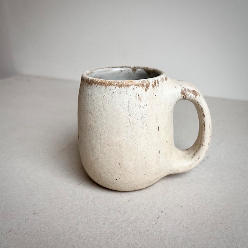 May include: A white ceramic mug with a rounded shape and a handle. The mug has a rustic, slightly distressed finish.