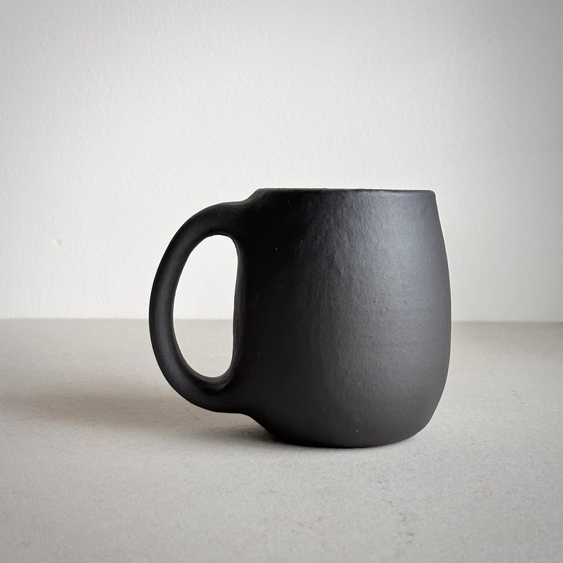May include: A black ceramic mug with a rounded body and a handle. The mug is made of a smooth, matte finish.