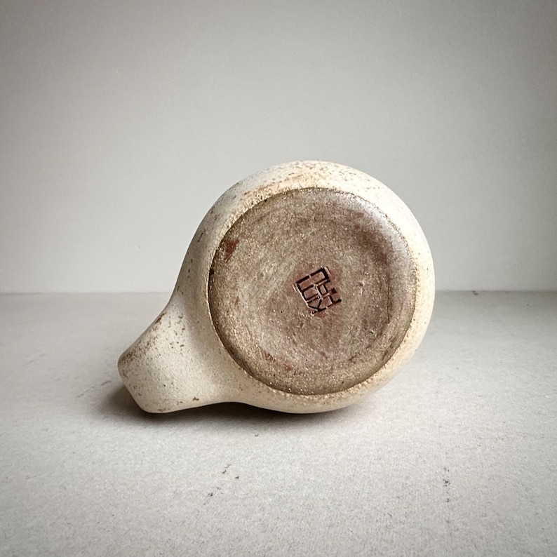 May include: A white ceramic pitcher with a brown glaze on the bottom. The pitcher has a small, square, brown stamp on the bottom that reads "田口".