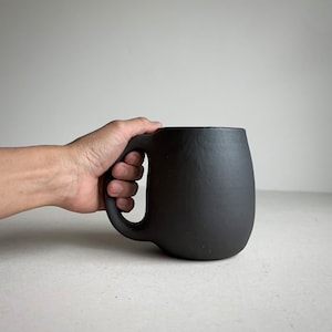 May include: A large, black ceramic mug with a rounded body and a thick handle. The mug has a textured, slightly rough surface.