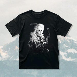 May include: A black short-sleeved t-shirt featuring a high-contrast, black and white photographic print of a person with voluminous blonde hair wearing a dark, embellished jacket with a wide collar and intricate detailing on the front chest area.
