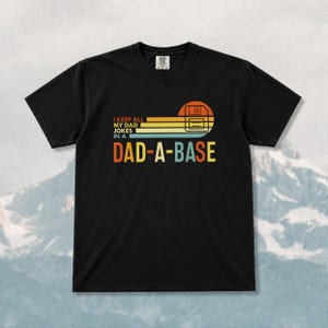 May include: Black t-shirt featuring the text "I KEEP ALL MY DAD JOKES IN A DAD-A-BASE" in orange, yellow, and blue. The design includes a retro-style graphic of a floppy disk and a sunset.