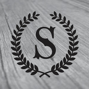 May include: A black monogram letter "S" inside a laurel wreath on a gray wood grain background. The wreath is composed of detailed leaves, and the letter has an elegant, stylized design. The overall aesthetic is classic and decorative.