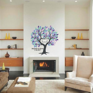 May include: A wall decal of a tree with purple, blue, and green leaves. The tree has a heart-shaped cutout in the center and the names "Manuela & Steven" with the date "9-10-2017" below. The decal is on a white wall above a fireplace.