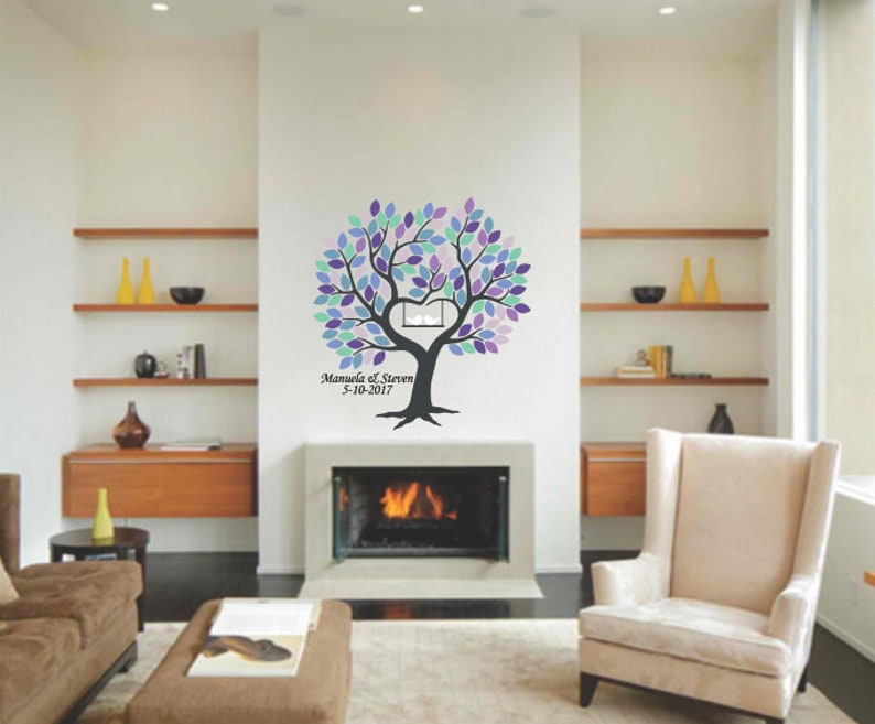 May include: A wall decal of a tree with purple, blue, and green leaves. The tree has a heart-shaped cutout in the center and the names "Manuela & Steven" with the date "9-10-2017" below. The decal is on a white wall above a fireplace.