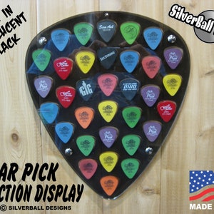 May include: Translucent black guitar pick collection display. The display is shaped like a guitar pick and holds numerous colorful guitar picks. The text "Guitar Pick Collection Display" is visible. The display is mounted on a wooden surface.