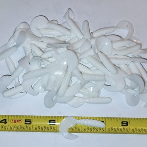 May include: A pile of white, soft plastic fishing lures with a curly tail design. A yellow measuring tape is in the foreground, showing the lure is approximately 4 inches long. The lures are designed to attract fish.