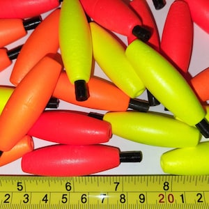 May include: A close-up of a pile of brightly colored fishing bobbers. The bobbers are yellow, orange, and red. A measuring tape is visible at the bottom of the image with the numbers 1 through 9.