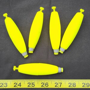 May include: Six bright yellow fishing bobbers with silver tops. The bobbers are arranged in a semi-circle on a dark gray surface.