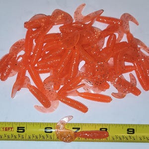 May include: A pile of orange and silver glitter soft plastic fishing lures. The lures are shaped like shrimp and are about 2 inches long. A measuring tape is in the bottom of the image with the numbers 4, 5, 6, 7, 8, 9, and 10 visible.