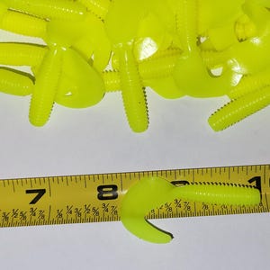 May include: A close-up of a pile of bright yellow plastic fishing lures. The lures are about 2 inches long and have a ribbed body with a curved tail. A ruler is in the bottom of the image to show the size of the lures.