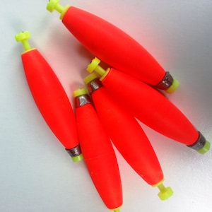 May include: Five bright orange bobbers with yellow tops and silver weights. The bobbers are arranged in a cluster on a white surface.