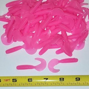 May include: A pile of bright pink, soft plastic fishing lures with a curly tail design. The lures are arranged on a white surface next to a yellow measuring tape. The lures are approximately 3 inches long.