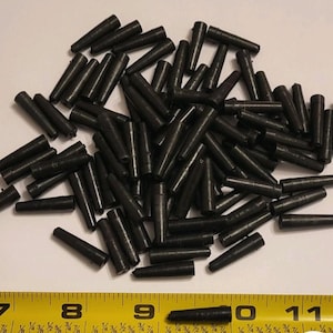 May include: A pile of small, dark gray cylindrical objects. The objects are scattered across a white surface, with a yellow measuring tape in the foreground. The objects appear to be approximately 1 inch long.