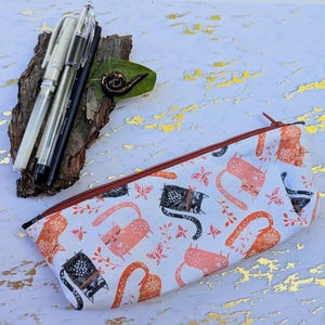 Op de afbeelding: Een witte etui met een herhaald patroon van oranje en zwarte katten, vlinders en bladeren. De etui heeft een bruine rits en bevat verschillende pennen. De achtergrond is een wit en goud oppervlak.