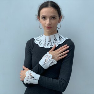 May include: A woman wearing a gray long-sleeved shirt with a white crocheted collar and matching cuffs. The collar and cuffs have a delicate, intricate design.