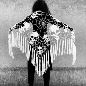 May include: A white crochet shawl with a spiderweb design and skull motifs. The shawl has fringe along the bottom edge.