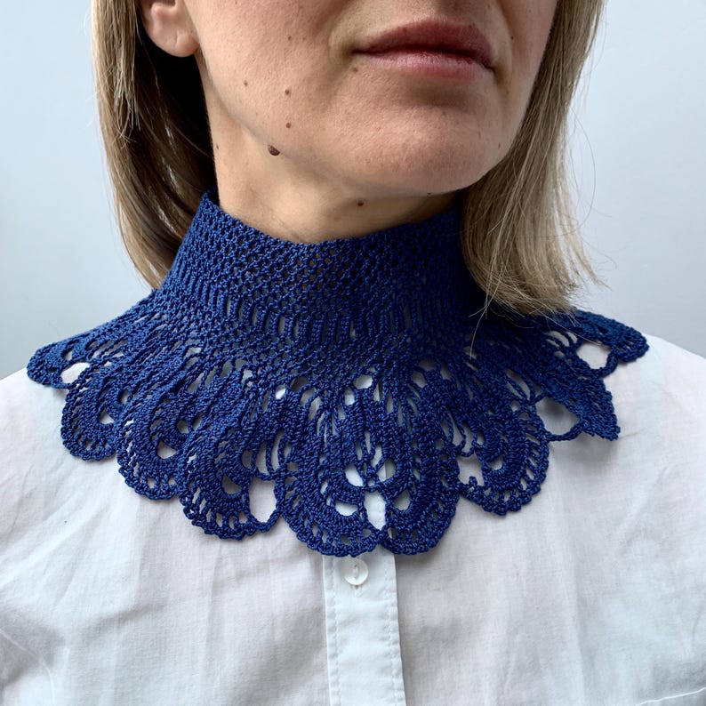Puede incluir: Un collar de ganchillo azul con un delicado patr&oacute;n de encaje. El collar se lleva alrededor del cuello y tiene un borde festoneado.