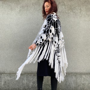 Pu&ograve; includere: Scialle in crochet bianco con un design a forma di teschio e frange. Lo scialle &egrave; drappeggiato sulle spalle di una persona e viene indossato con un abito nero.